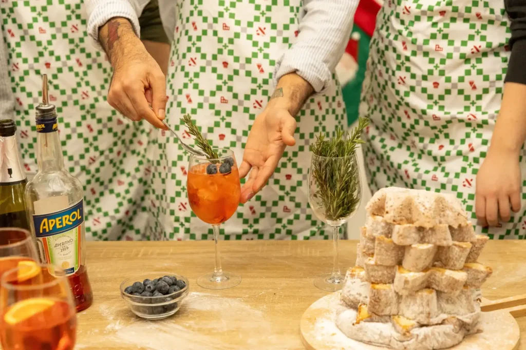 Close-up of hands garnishing orange cocktails with fresh rosemary and blueberries next to a decorative dessert tower during a culinary class