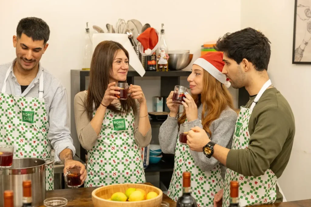 Four cooking class participants in festive attire enjoying hot mulled wine together with bowl of fresh citrus fruits on counter