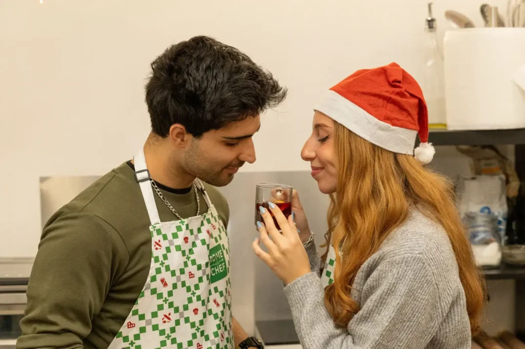 Young couple in festive aprons sharing a glass of homemade mulled wine in an intimate moment during Christmas cooking workshop