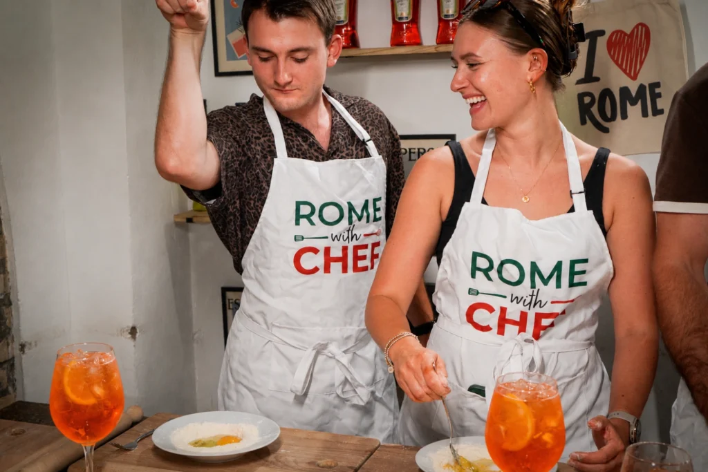 Two guests wearing Rome With Chef aprons laughing while making pasta alongside Aperol Spritz cocktails during a Carpe Diem Tours Rome spritz and spaghetti cooking class